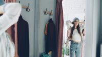 A woman in her 40s smiles as she tries on a pair of mid-rise, straight-leg jeans in a well-lit clothing store, checking the fit in a mirror.