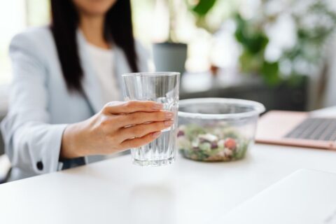 A busy professional sitting at a desk with a laptop, taking a sip of water from a glass bottle, with a bowl of berries and a banana nearby, symbolizing practical hydration during work.