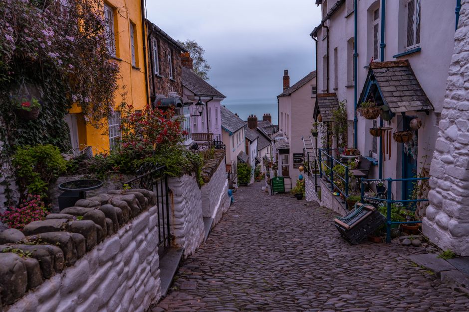 A picturesque, narrow cobblestone street in a quiet European coastal village, lined with colorful buildings and local flowers.