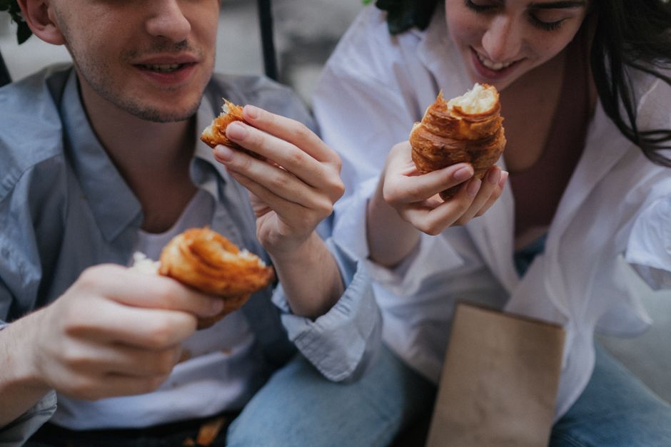 A couple enjoying freshly baked pastries and coffee at a cozy, charming bakery, with artisanal bread and treats visible in the background.