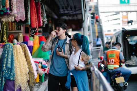 A couple happily browsing colorful fresh produce at a vibrant outdoor market, perhaps in Europe, with baskets and local goods on display.