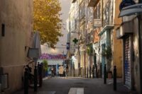 A couple strolls hand-in-hand down a narrow, sun-dappled cobblestone street in Marseille's Le Panier district, surrounded by ancient stone buildings.