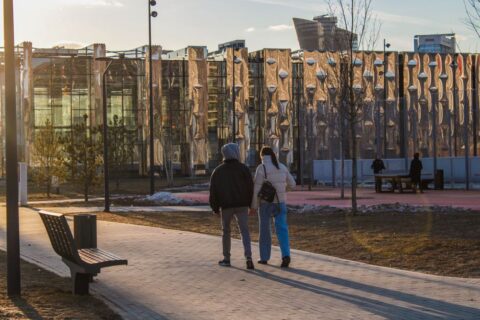 A couple walks hand-in-hand through a sun-drenched European city street during golden hour, long shadows stretching behind them.