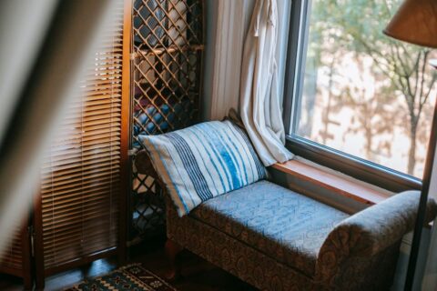 A cozy, minimalist reading corner in a small apartment, featuring a compact armchair, a tall, narrow bookshelf, and a soft blanket, illuminated by a floor lamp.