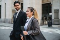 Three diverse professionals in smart casual attire walking purposefully down a tree-lined city street, one carrying a laptop bag.