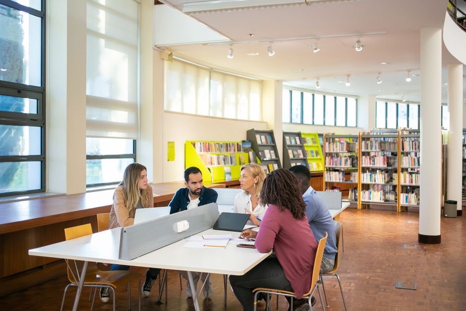 A diverse group of students from different backgrounds working together at a large table in a modern library, discussing and cross-referencing information.