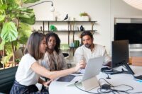 A small, diverse team of three people looking at a laptop screen, appearing to collaborate on a cybersecurity strategy, with a coffee cup and notebooks on the table.