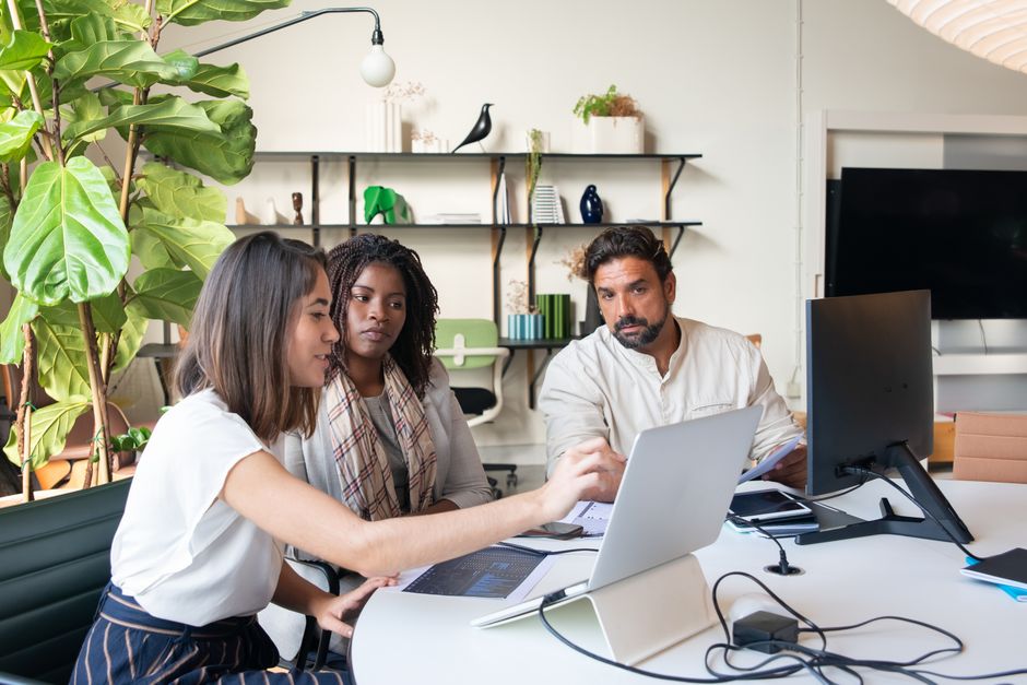 A small, diverse team of three people looking at a laptop screen, appearing to collaborate on a cybersecurity strategy, with a coffee cup and notebooks on the table.