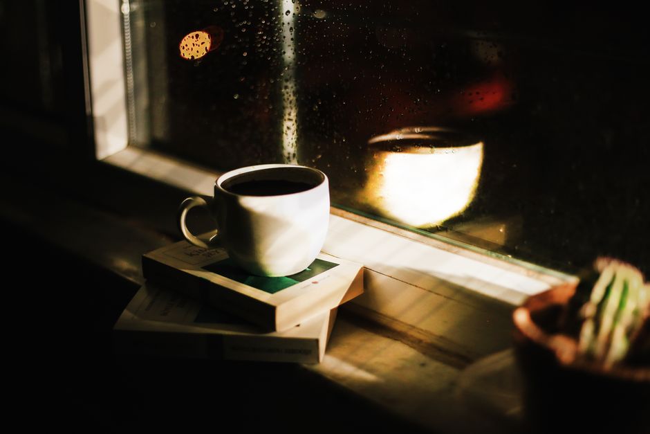 A steaming, half-empty coffee cup rests on a wooden window sill, with soft rain blurring the view outside, evoking a sense of calm and unhurried peace.