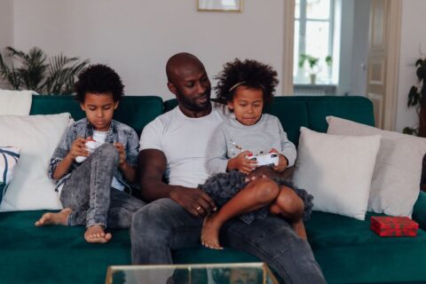 A family of four, including two children, laughing while playing a video game on a TV screen with two game controllers in hand.