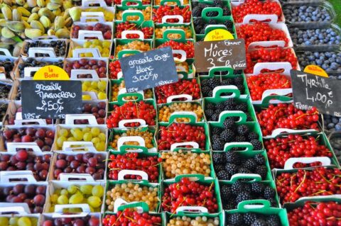 A bustling outdoor market in a French town, with stalls overflowing with colorful fresh fruits, vegetables, cheeses, and baked goods, under an awning.