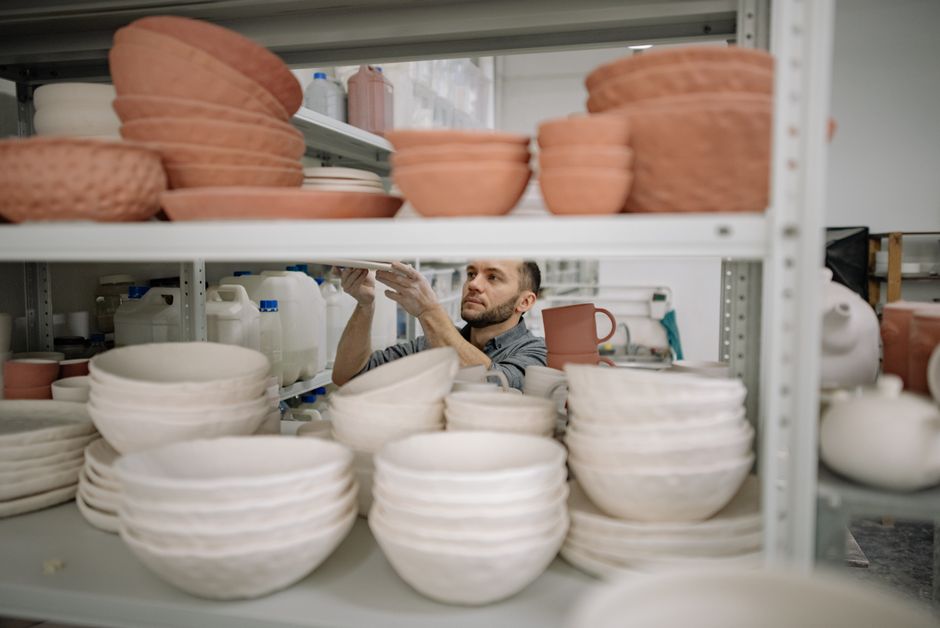 Close-up of hands thoughtfully arranging a collection of unique, handmade ceramic mugs and bowls on a rustic wooden shelf, emphasizing the curation of personal items.