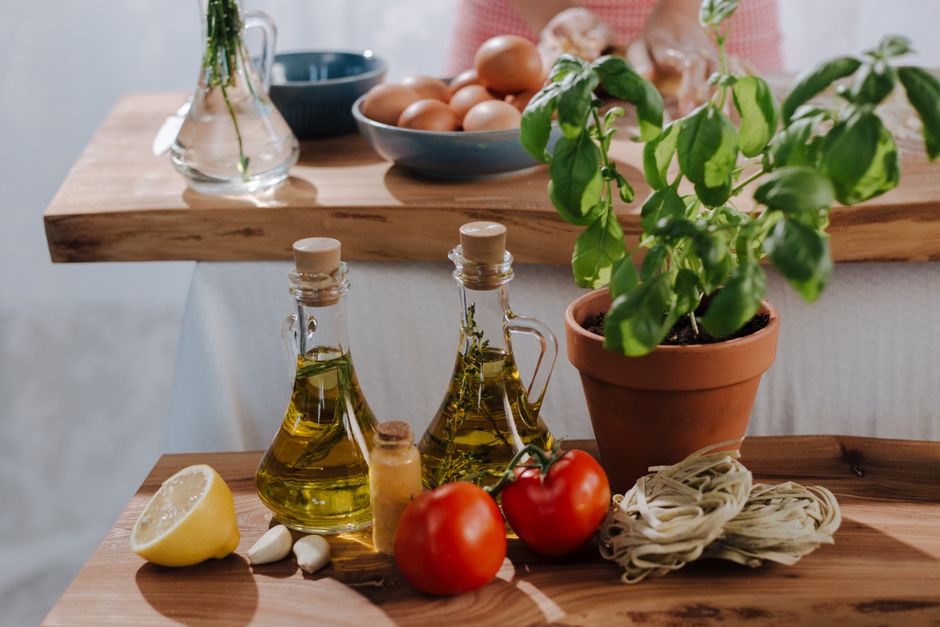 A collection of Mediterranean pantry staples on a wooden counter, including extra virgin olive oil, canned chickpeas, diced tomatoes, whole wheat pasta, and dried oregano.