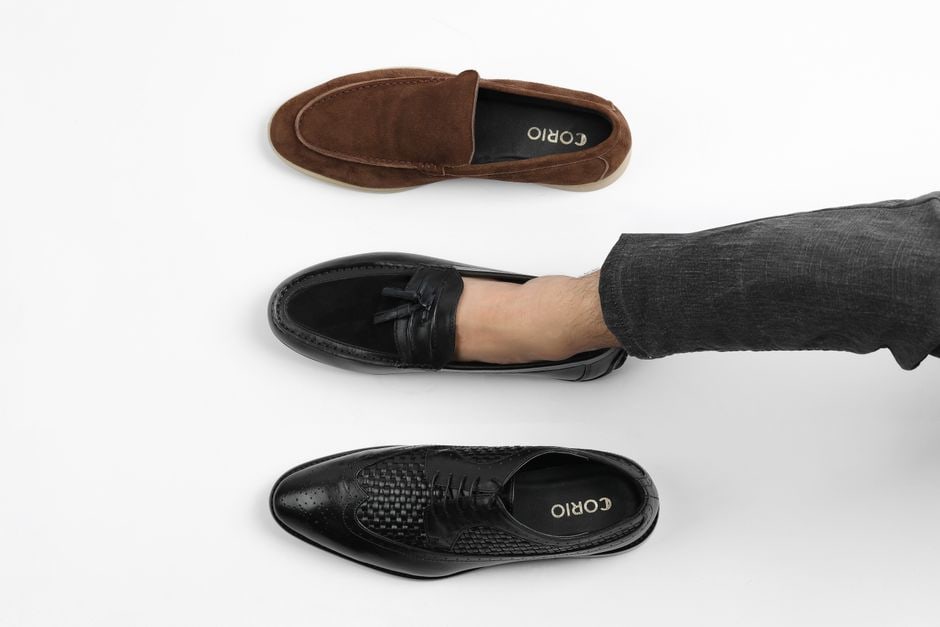 A man in tailored trousers and a collared shirt, sitting on a low wall in a city park, putting on a pair of sleek leather loafers, having just removed his casual walking shoes.