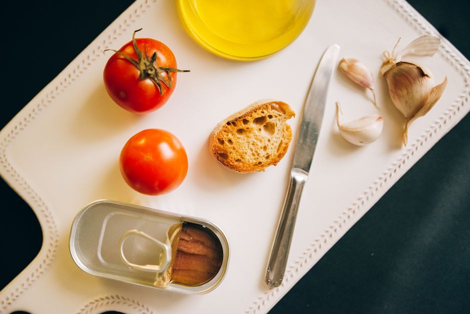 An overhead shot of a wooden table covered with a diverse array of fresh, colorful produce including leafy greens, bell peppers, tomatoes, herbs, olive oil, and a baguette, symbolizing a market basket meal.