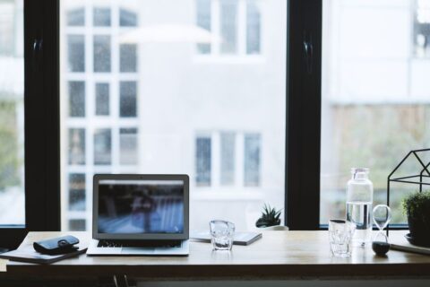 A clean, minimalist home office space with a wooden desk, a laptop, a plant, and a comfortable chair, bathed in natural light.