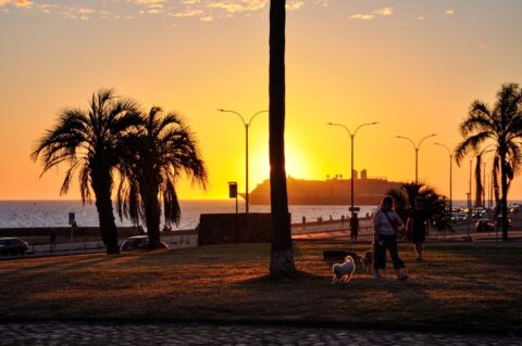 A couple watches the sunset over the Rio de la Plata from the Montevideo Rambla, with the city skyline softly lit in the background.