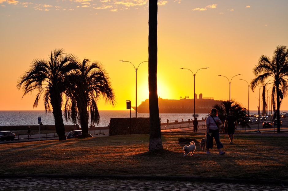 A couple watches the sunset over the Rio de la Plata from the Montevideo Rambla, with the city skyline softly lit in the background.