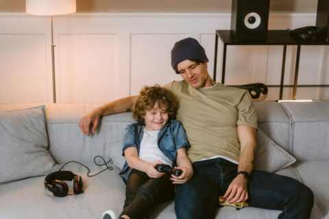 A parent and child sitting together on a sofa, the child absorbed in a video game while the parent gently observes and converses with them.