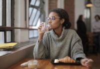 A person in a business casual attire sipping coffee and eating a pastry at a bustling European cafe, looking at a laptop.