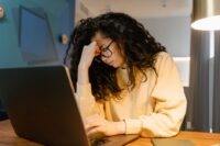 A person with a strained expression covering their face, sitting in front of a laptop with multiple video call windows open, illustrating video call fatigue.