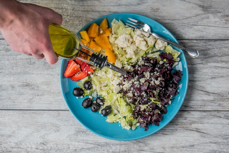 A close-up shot of a hand drizzling extra virgin olive oil over a fresh, green salad in a bowl, illustrating the role of healthy fats in a hydrating diet.