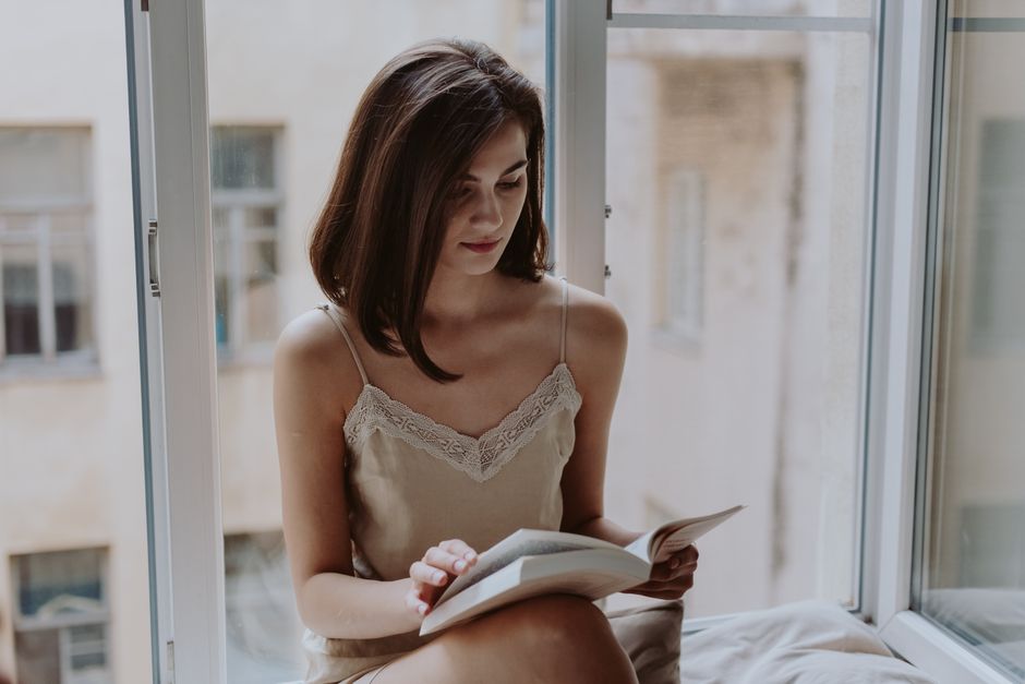 A person reading a physical book under a warm lamp in a comfortable living room, with a soft throw blanket and a plant, indicating evening relaxation.