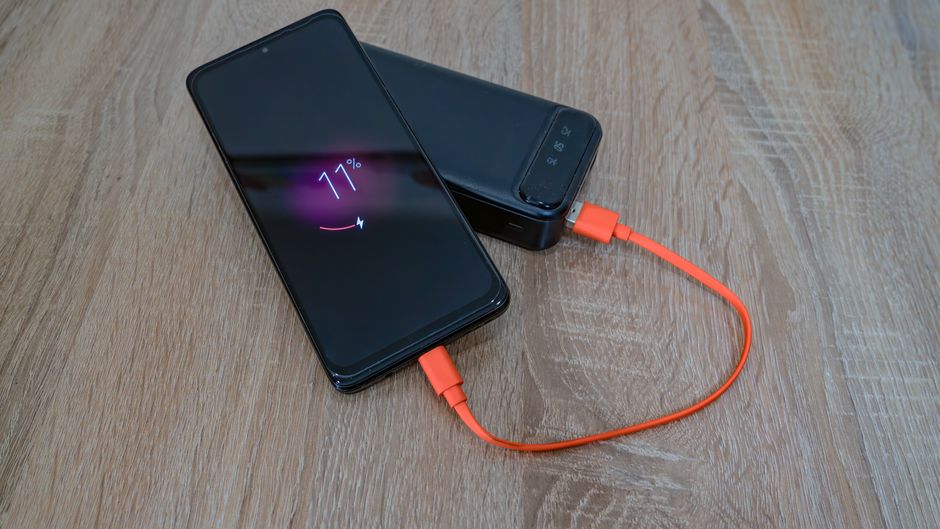 A portable power bank charging a smartphone and tablet on a wooden table, with travel items in the background.
