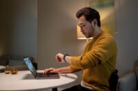 A remote worker in a bright home office, looking thoughtfully at their smartwatch, with a laptop in the background.