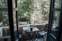 A neatly arranged small balcony with a foldable bistro table, two chairs, and several potted plants, inviting a morning coffee ritual.