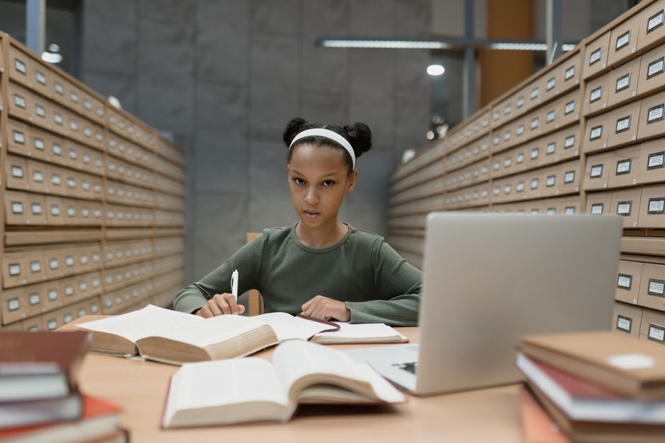 A student in a library setting, looking thoughtfully at a laptop screen with books open around them, symbolizing the research process with AI tools.