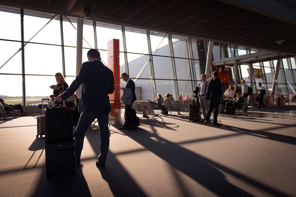 A person in smart business casual attire (blazer, dark trousers, stylish walking shoes) navigating a European city airport with a single carry-on bag.