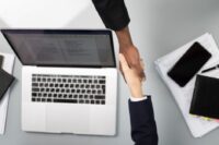 Two business partners shaking hands across a table with a laptop and papers, signifying a new agreement.