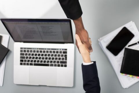 Two business partners shaking hands across a table with a laptop and papers, signifying a new agreement.