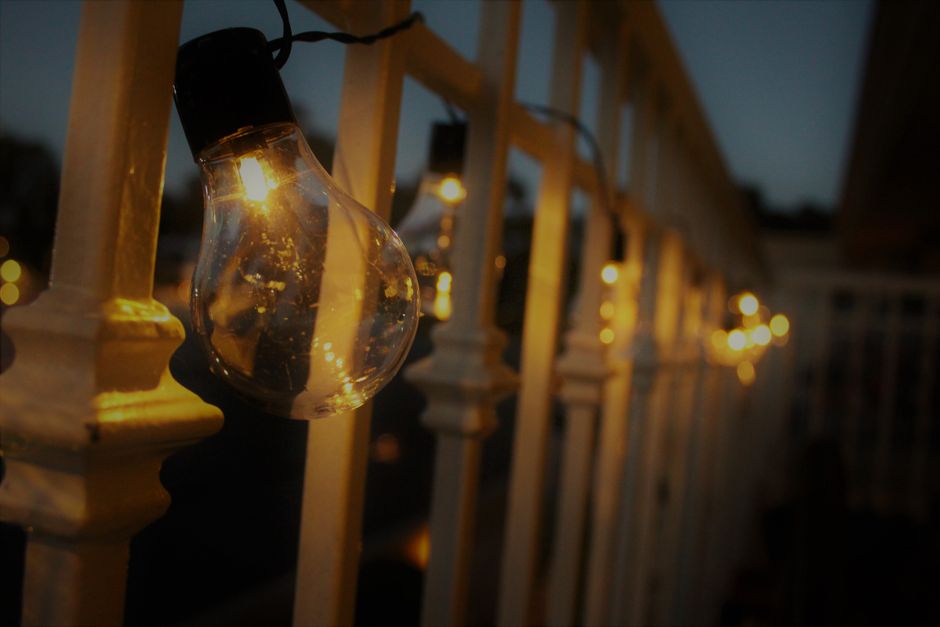 An urban balcony at dusk, illuminated by warm string lights, featuring a railing planter filled with flowers and a comfortable throw on a chair, perfect for evening air.