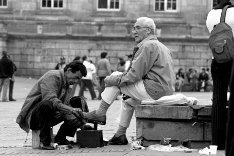 A woman sitting on a bench in a European city square, swapping her comfortable walking sneakers for elegant ballet flats, with a small handbag beside her.