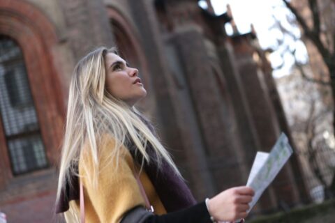 A young woman, smiling and holding a map, standing on a charming cobblestone street in a European-style city, looking curious and ready to explore.