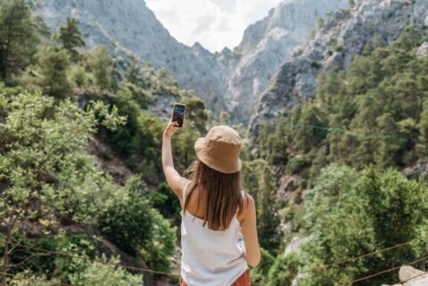 A woman backpacker checks her phone, likely reviewing photos, with a stunning mountain landscape in the background, illustrating the travel context of photo backup.