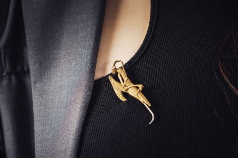 Woman's hand carefully pinning an ornate vintage brooch to the lapel of a gray wool blazer, showing how brooches add detail.
