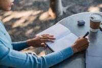 A person with dark skin writes in a leather-bound travel journal at a rustic wooden table in a sunlit café, a half-empty coffee cup beside them, illustrating thoughtful reflection during a trip.